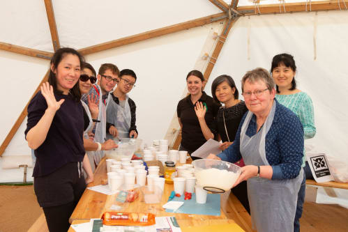 Eight people in a community group smiling and waving. They are standing around a wooden table in a white tent. Their are baking ingredients on the table. The person in the front right holds a bowl and stirs ingredients.