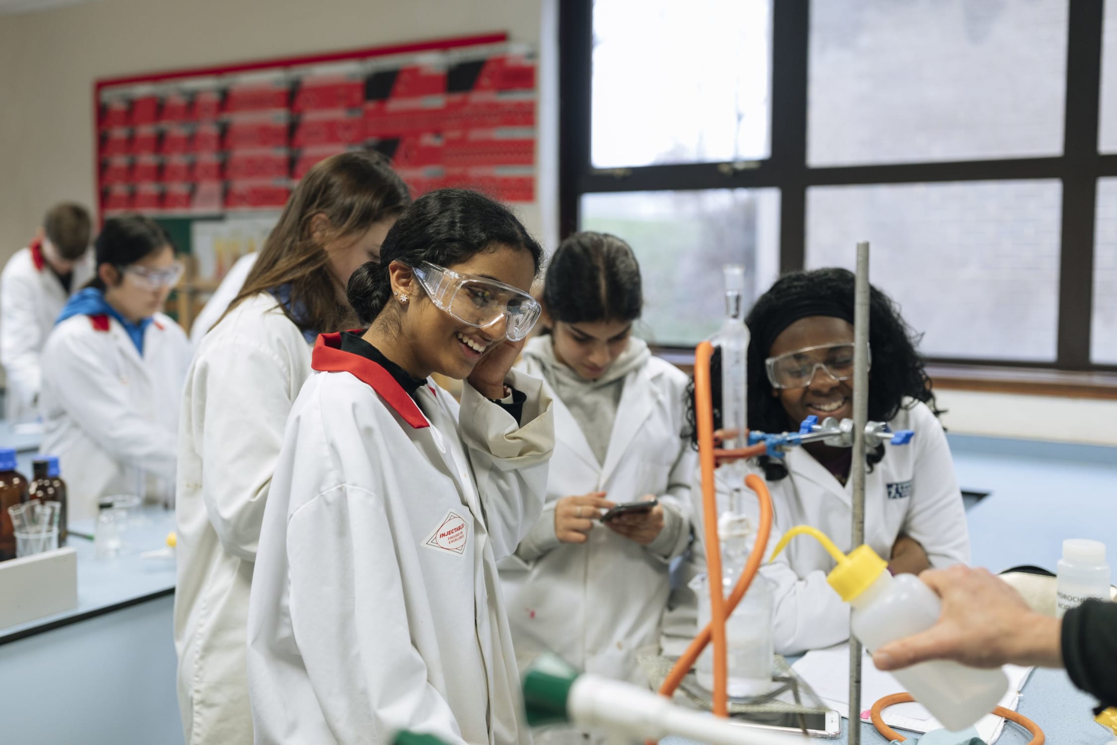 Two girls in a classroom, smiling, wearing white lab coats and goggles, conducting an experiment
