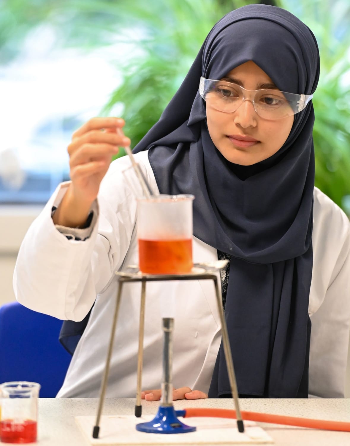 Girl wearing hijab, white lab coat and goggle in a classroom, adding liquid to a beaker